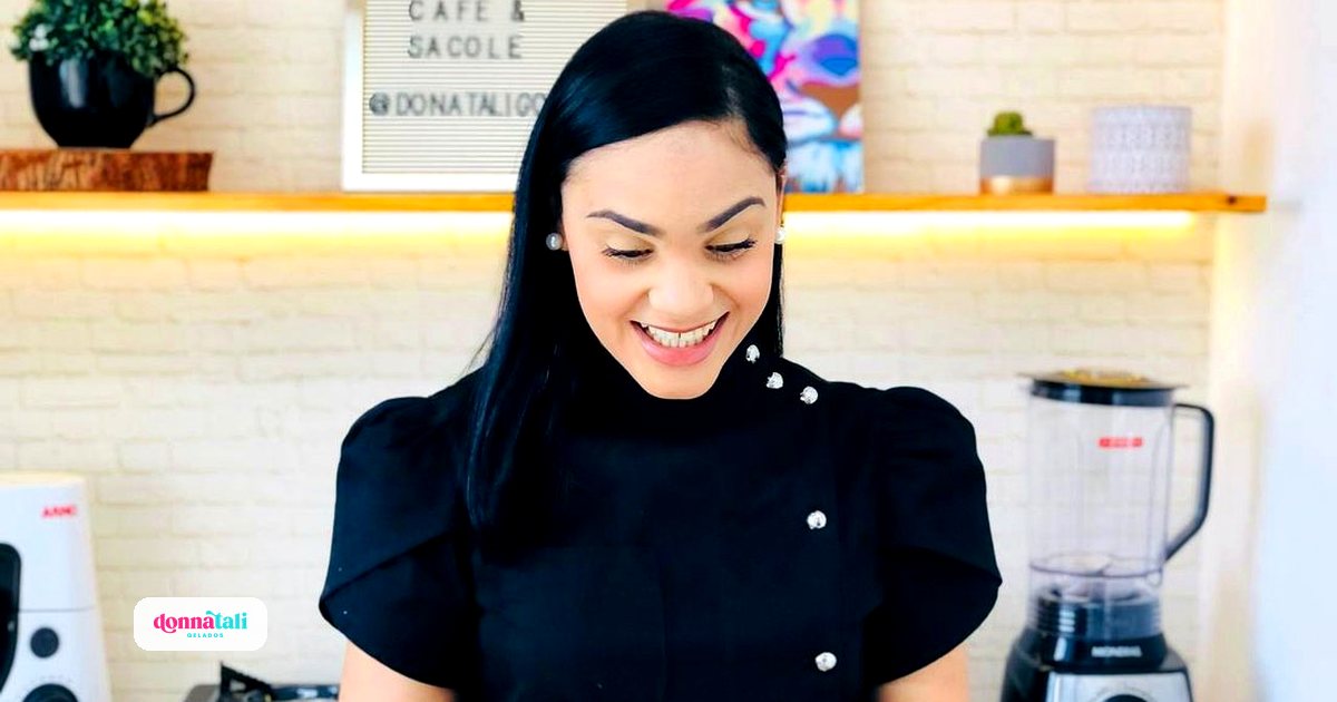 Talita, da Donnatali Gelados, sorrindo no seu espaço de trabalho simples, com liquidificador e fogão portátil ao fundo